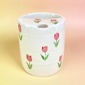 May include: A white ceramic vase with a speckled finish, adorned with hand-painted pink tulips and green leaves. The vase has a curved top with a hole and is cylindrical in shape. The floral design adds a touch of spring to any room.