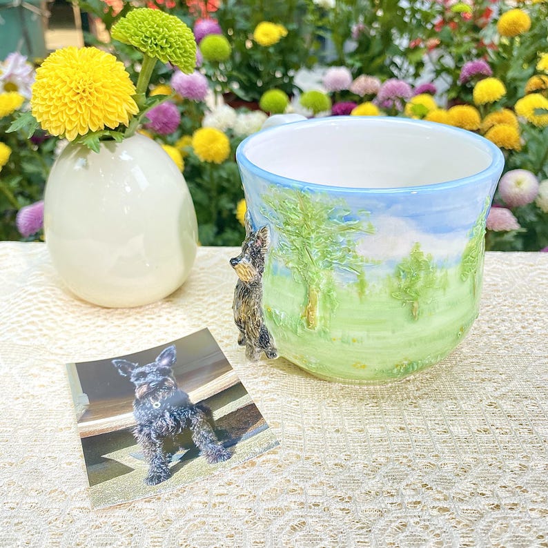 May include: A ceramic mug with a landscape design featuring a dog figurine handle. The mug has a light blue sky and green grass design. A small white vase with yellow and green flowers is next to the mug. A photo of a dog is in front of the mug.