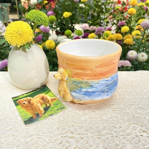 May include: A ceramic mug with a golden retriever dog sculpture on the side. The mug features a painted sunset scene with orange, blue, and yellow hues. A small photo of a golden retriever is next to the mug. A white vase with yellow flowers is also in the image.