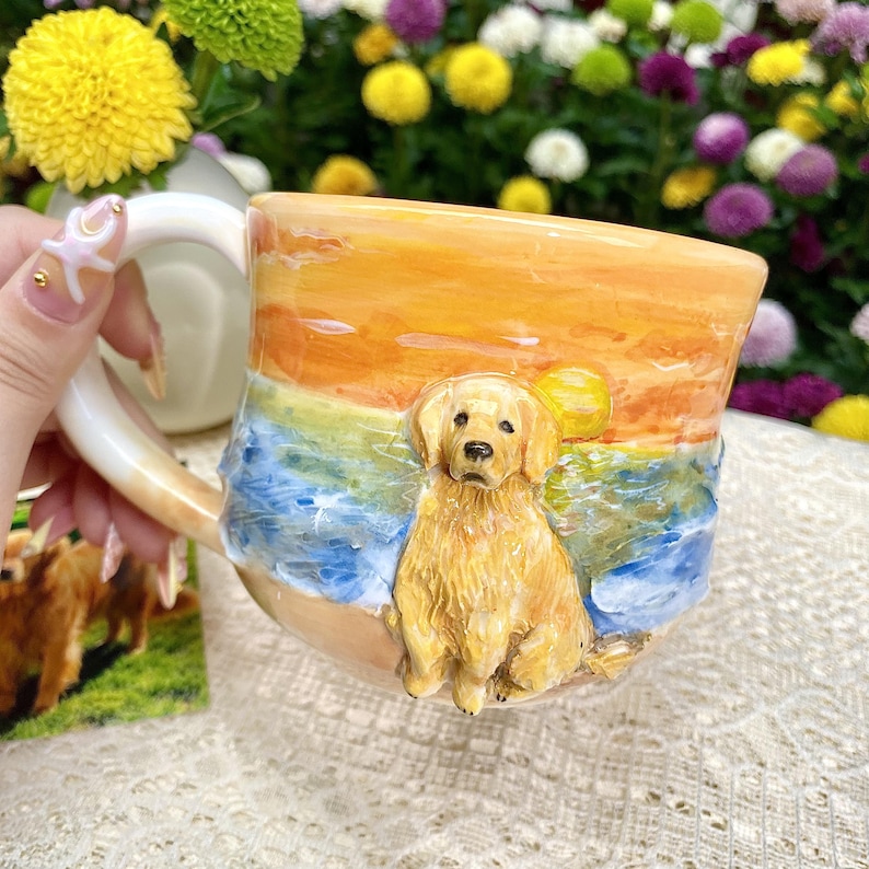 May include: A ceramic mug featuring a golden retriever on a beach scene with a sunset backdrop. The mug has a curved handle and is painted with shades of orange, yellow, and blue. The dog is sculpted in relief, adding a 3D effect.