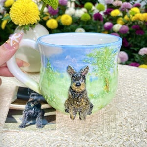 May include: A ceramic mug with a hand-painted landscape scene featuring a black and brown dog. The mug has a white handle and a blue and green background. A small photo of the dog is next to the mug.