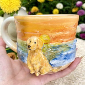 May include: A ceramic mug featuring a golden retriever dog sculpture. The mug has a painted sunset scene with shades of orange, yellow, and blue. The mug has a curved handle and is held in a person's hand.