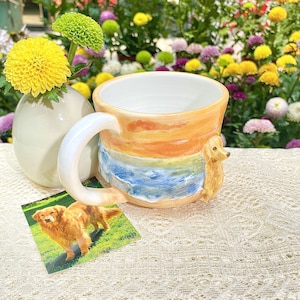 May include: A ceramic mug with a sunset and ocean design, featuring a golden retriever figurine on the side. The mug has a white interior and a large handle. A small photo of a golden retriever is next to the mug.