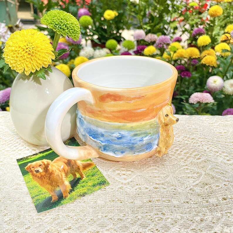 May include: A ceramic mug with a sunset and ocean design, featuring a golden retriever figurine on the side. The mug has a white interior and a large handle. A small photo of a golden retriever is next to the mug.