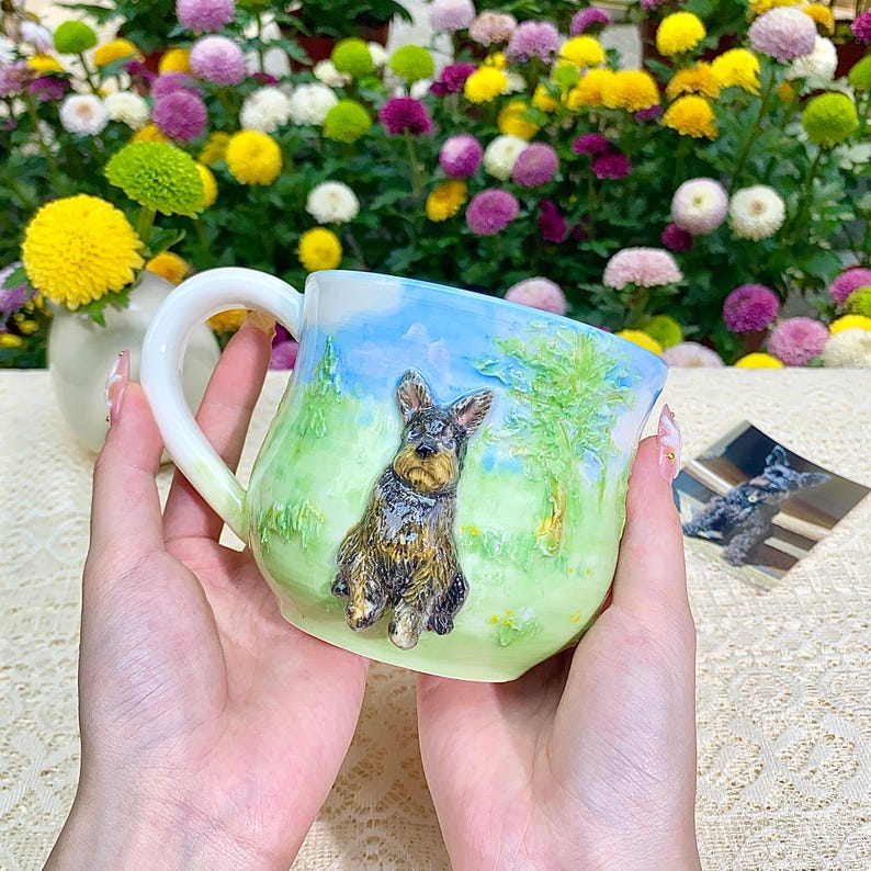 May include: A ceramic mug with a detailed painting of a dog on a green and blue landscape. The mug has a white handle and is held in a person's hands. A small photo of the dog is visible.