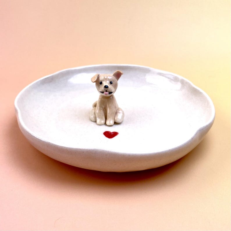 Peut inclure: Un plat en c&eacute;ramique blanc avec un bord ondul&eacute;, orn&eacute; d'une figurine de chien beige. Un c&oelig;ur rouge est peint sous le chien. Le plat est pos&eacute; sur un fond de couleur p&ecirc;che clair.
