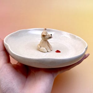 Peut inclure: Un plat en c&eacute;ramique cr&egrave;me avec une figurine de chien assis et un c&oelig;ur rouge. Le plat a un bord l&eacute;g&egrave;rement ondul&eacute;. Le chien est brun clair avec des yeux noirs et une langue rose. Le c&oelig;ur est une petite forme rouge.