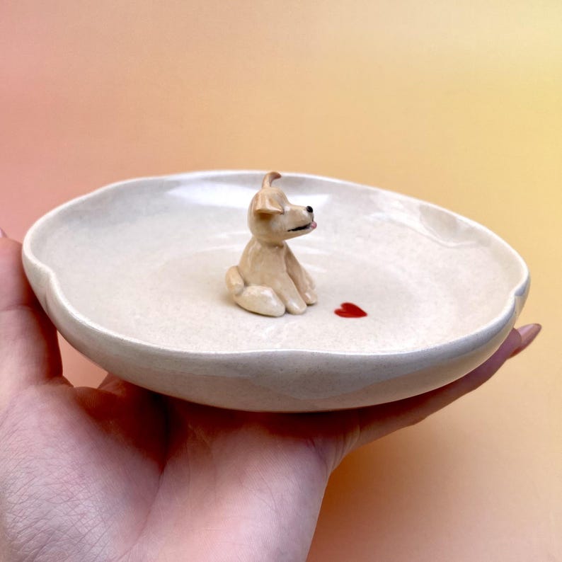 Peut inclure: Un plat en c&eacute;ramique cr&egrave;me avec une figurine de chien assis et un c&oelig;ur rouge. Le plat a un bord l&eacute;g&egrave;rement ondul&eacute;. Le chien est brun clair avec des yeux noirs et une langue rose. Le c&oelig;ur est une petite forme rouge.