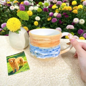 May include: A ceramic mug with a beach-themed design. The mug features a light tan top, blue waves, and a white interior. A small photo of a golden retriever is next to the mug. A small vase with yellow flowers is also in the image.