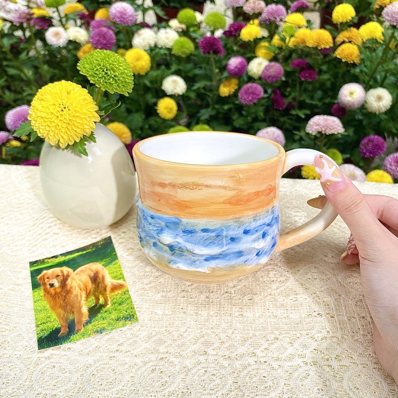 May include: A ceramic mug with a beach-themed design. The mug features a light tan top, blue waves, and a white interior. A small photo of a golden retriever is next to the mug. A small vase with yellow flowers is also in the image.