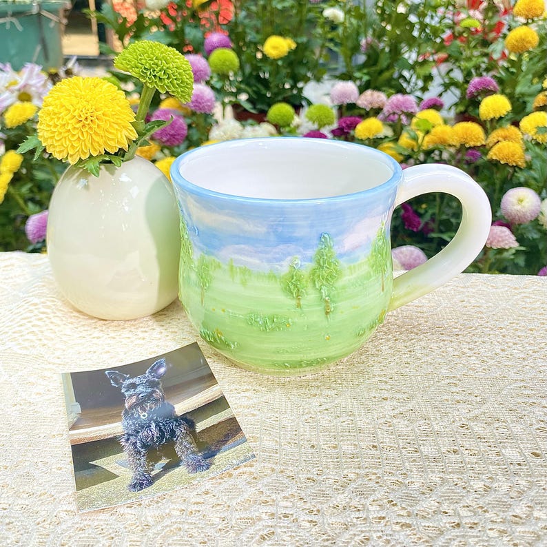 May include: A handmade ceramic mug with a landscape design featuring a blue sky, green grass, and trees. A small white vase holds yellow and green flowers. A photo of a dog is in the foreground.