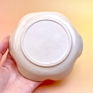 Peut inclure: Un bol en c&eacute;ramique cr&egrave;me avec un bord festonn&eacute;, tenu dans une main. Le dessous du bol est visible, avec une base circulaire l&eacute;g&egrave;rement textur&eacute;e. Le design du bol est simple et &eacute;l&eacute;gant, adapt&eacute; au service ou &agrave; l'exposition.