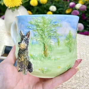 May include: A ceramic mug with a sculpted Yorkshire Terrier dog on the side. The mug features a landscape design with a blue sky, green grass, and trees. The mug is hand-painted with vibrant colors.