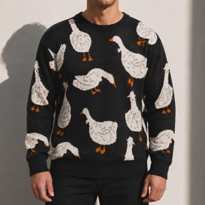 May include: A black long-sleeve crewneck sweater featuring a repeating pattern of white geese with orange beaks and feet. The knit garment has a ribbed neckline, cuffs, and hem, showcasing multiple geese in various poses across the front and sleeves.
