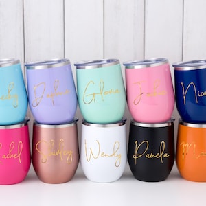 Puede incluir: Una colección de vasos de vino sin pie en varios colores, incluyendo azul, morado, verde, rosa y naranja. Cada vaso tiene un nombre en oro. Los vasos se exhiben sobre una superficie blanca.