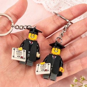 Pode incluir: Dois chaveiros com tema de formatura com figuras estilo Lego. Cada figura usa uma beca e um capelo pretos, segurando um diploma. Os chaveiros têm argolas e correntes de metal prateado.