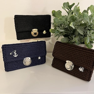 Op de afbeelding: Drie gehaakte clutch portemonnees in zwart, marineblauw en bruin. Elke portemonnee heeft een zilveren sluiting en decoratieve versieringen. De zwarte portemonnee heeft een gouden sluiting en een kleine gouden kraal. De marineblauwe portemonnee heeft een anker speld en een ster. De bruine portemonnee heeft een vlinder.