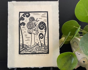 May include: Black and white linocut print of a snail on a mushroom with fern-like plants in the background. The print is signed "Ride Along 740 VE Oakley, January 2022".
