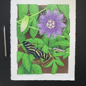 Puede incluir: Una impresión en madera de una flor de la pasión púrpura, una mariposa zebra de alas largas y una oruga en una vid. La impresión se titula "Passion Vine and Zebra Longwing" y está firmada por la artista, "Wendy Hollingsworth".