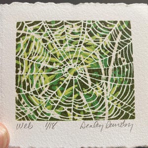 May include: A small, square print of a spider web in shades of green and white. The artwork is on textured, off-white paper. The word "Web" and the number "4/18" are printed at the bottom left. The artist's signature is at the bottom right.