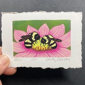 Puede incluir: Una impresión en madera de dos abejorros en una flor rosa. La impresión se titula "Bees" y está numerada 1/20. La firma del artista, "Dealey Deustch", está en la esquina inferior derecha.