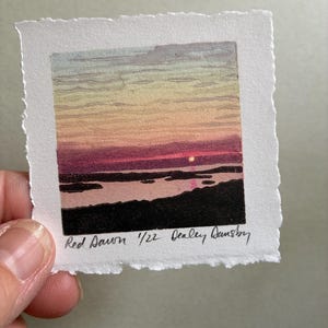 May include: A small square print on textured white paper featuring a sunrise landscape with a dark foreground, a pink and purple horizon, and a pale yellow sky. The text 'Red Dawn 1/22 Dealey Dansby' is handwritten in black ink at the bottom.