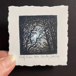 Peut inclure: Une estampe en noir et blanc d'une pleine lune brillant à travers les branches d'arbres. L'estampe est intitulée "Wolf Moon 4/20" et signée "Audrey Daustry".