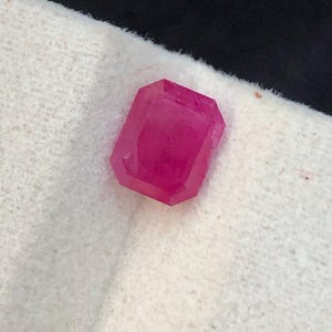 May include: A faceted, rectangular ruby gemstone with a deep pink hue. The gem is set against a white, textured background, with a dark backdrop. The cut of the stone allows light to pass through, highlighting its clarity and color.