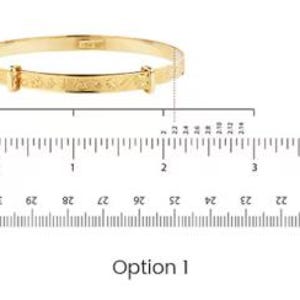 May include: A gold-colored adjustable baby bangle bracelet. The bracelet is shown with a ruler, measuring approximately 2 inches in diameter. The bracelet has a decorative engraved design and a clasp closure. The text "Option 1" is below the bracelet.