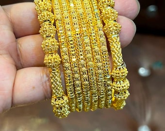 May include: A set of seven gold-colored bangles featuring a metallic finish. The two outer bangles display a prominent beaded and textured pattern, while the five inner bangles exhibit a consistent, repeating geometric engraved design.