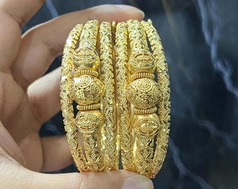 May include: Close-up of several gold bangles with intricate, ornate designs. The bangles have a textured surface and are held in a hand against a dark background. The bangles are a bright, shiny gold color.