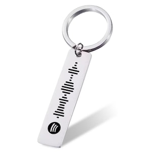 May include: A silver-colored keychain with a rectangular tag featuring a black soundwave pattern and a Spotify logo. The keychain has a circular ring for attaching keys.