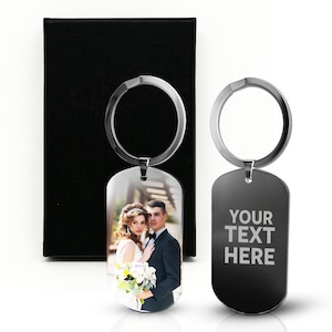 May include: Two metal keychains with silver rings. One keychain has a photo of a couple in formal attire. The other keychain is black with the text "YOUR TEXT HERE" in white. A black gift box is in the background.