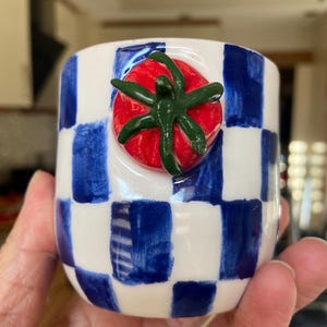 May include: A ceramic cup with a blue and white chequered pattern. A red tomato with green leaves is affixed to the front of the cup. The cup is held in a person's hand.