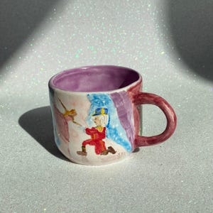 Może przedstawiać: Ceramiczny kubek z malowaną ilustracją żołnierza w czerwonym mundurze i mieczem. Kubek ma fioletowe wnętrze, bordowy uchwyt i kolorową powierzchnię zewnętrzną z odcieniami niebieskiego, różowego i fioletowego.