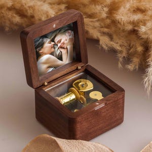 Könnte beinhalten: Eine Holz-Spieluhr mit einem Klappdeckel, die ein gerahmtes Foto eines Paares zeigt. Die Box ist geöffnet und zeigt einen goldfarbenen Musikmechanismus und ein glasbedecktes Fach. Die Box ist aus dunklem Holz.