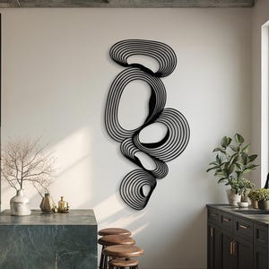 May include: Black metal wall art with a layered, abstract design. The artwork features a series of interconnected, flowing shapes composed of parallel lines, creating a three-dimensional effect. The piece is mounted on a white wall in a modern kitchen setting.