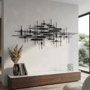 Könnte beinhalten: Eine Wandskulptur aus schwarzem Metall mit einer geometrischen Anordnung aus horizontalen und vertikalen Stäben unterschiedlicher Länge. Die abstrakte Komposition ist an einer hellen Wand über einem niedrigen Holzschrank mit weißer Marmorplatte montiert.