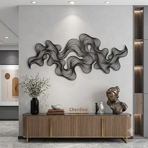 Könnte beinhalten: Ein Wandkunstobjekt aus schwarzem Metall, das aus einer Reihe miteinander verbundener, fließender, wellenförmiger Linien besteht und eine abstrakte, wolkenartige Form bildet. Es ist horizontal an einer hellgrauen Wand über einem Holzschrank montiert.