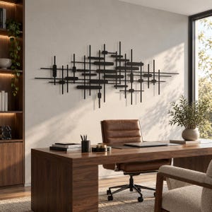 May include: A large black metal wall sculpture featuring a complex arrangement of intersecting horizontal and vertical bars and rectangular shapes, creating a geometric grid pattern mounted on a plain light-colored wall above a wooden desk.