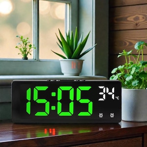 Puede incluir: Reloj despertador digital negro que muestra la hora 15:05 en verde, con una temperatura de 34°C. El reloj tiene dos iconos de alarma y está sobre una superficie de madera.