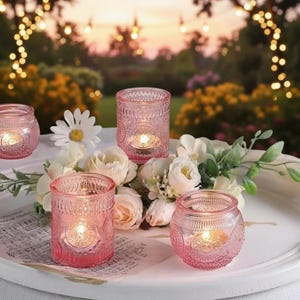 Puede incluir: Candelabros de vidrio rosa con intrincados patrones en relieve, cada uno con una vela encendida. Los candelabros están dispuestos en una bandeja blanca con flores blancas y vegetación, con un fondo borroso al aire libre.