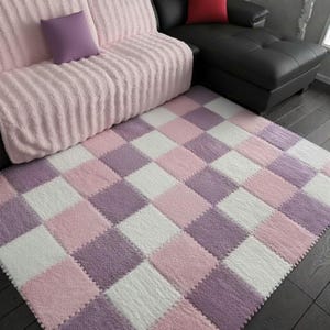 May include: A soft, plush rug with a checkerboard pattern in shades of pink, white, and purple. The rug is made of a textured material and is placed on a dark wood floor. The rug is near a pink couch and a black couch.