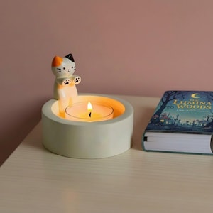 Puede incluir: Un portavelas en forma de gato verde claro con una vela de té encendida. La figura del gato tiene detalles en naranja, negro y blanco. Un libro titulado "The Lumina Woods" está al lado del portavelas.