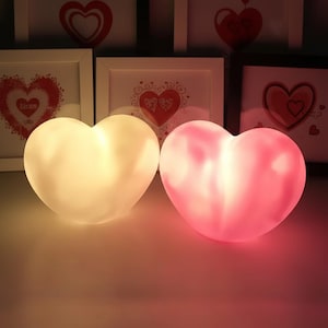 May include: Two heart-shaped LED lights, one glowing warm white and the other pink, sit side-by-side. The lights are on a reflective surface, with framed heart art in the background. The lights are a romantic decorative item.