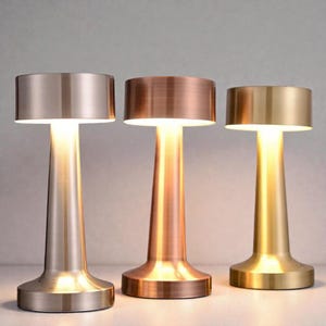 May include: Three contemporary table lamps with metallic finishes. Each lamp features a cylindrical shade and a conical base. The lamps are in silver, copper, and gold. They are illuminated, giving off a warm light.