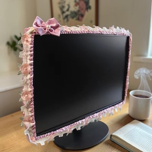 Pode incluir: Uma moldura decorativa para monitor com fita de cetim rosa e acabamento em renda. A moldura é adornada com pequenas rosas de tecido rosa e um laço de cetim com um detalhe de pérola. A moldura é colocada em um monitor de computador preto.