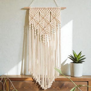 May include: Cream-colored macrame wall hanging with a geometric design and long fringe. The wall decor is suspended from a wooden dowel and hangs against a neutral wall. A small potted plant sits on a wooden surface below.