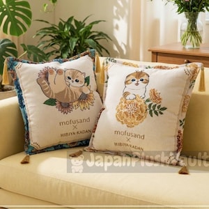 May include: Two decorative throw pillows with cat illustrations. One pillow features a cat hanging from a sunflower, the other shows a cat sitting on a flower. Both pillows have the text "mofusand X HIBIYA-KADAN". The pillows have tassels.