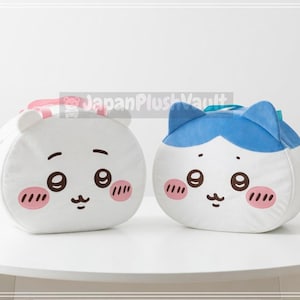 以下が含まれることがあります： 丸いキャラクターのぬいぐるみ枕が2つ。1つはピンクの頬、ピンクのリボン、クマの耳が付いた白。もう1つは青いアクセントと猫の耳が付いた白。どちらも黒い目と小さな口があります。
